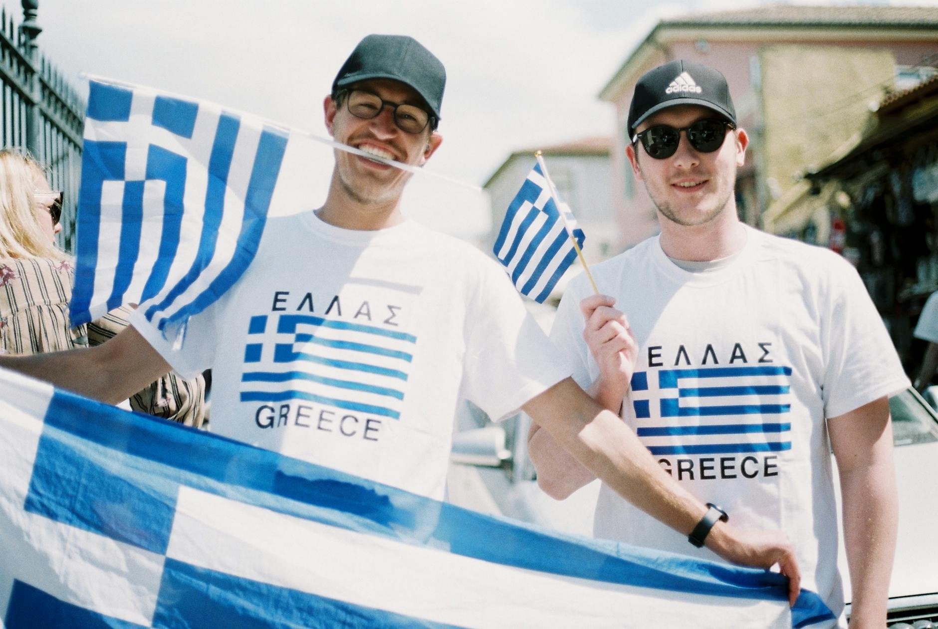 мъж, облечен с тениски и държащ знамена на Гърция